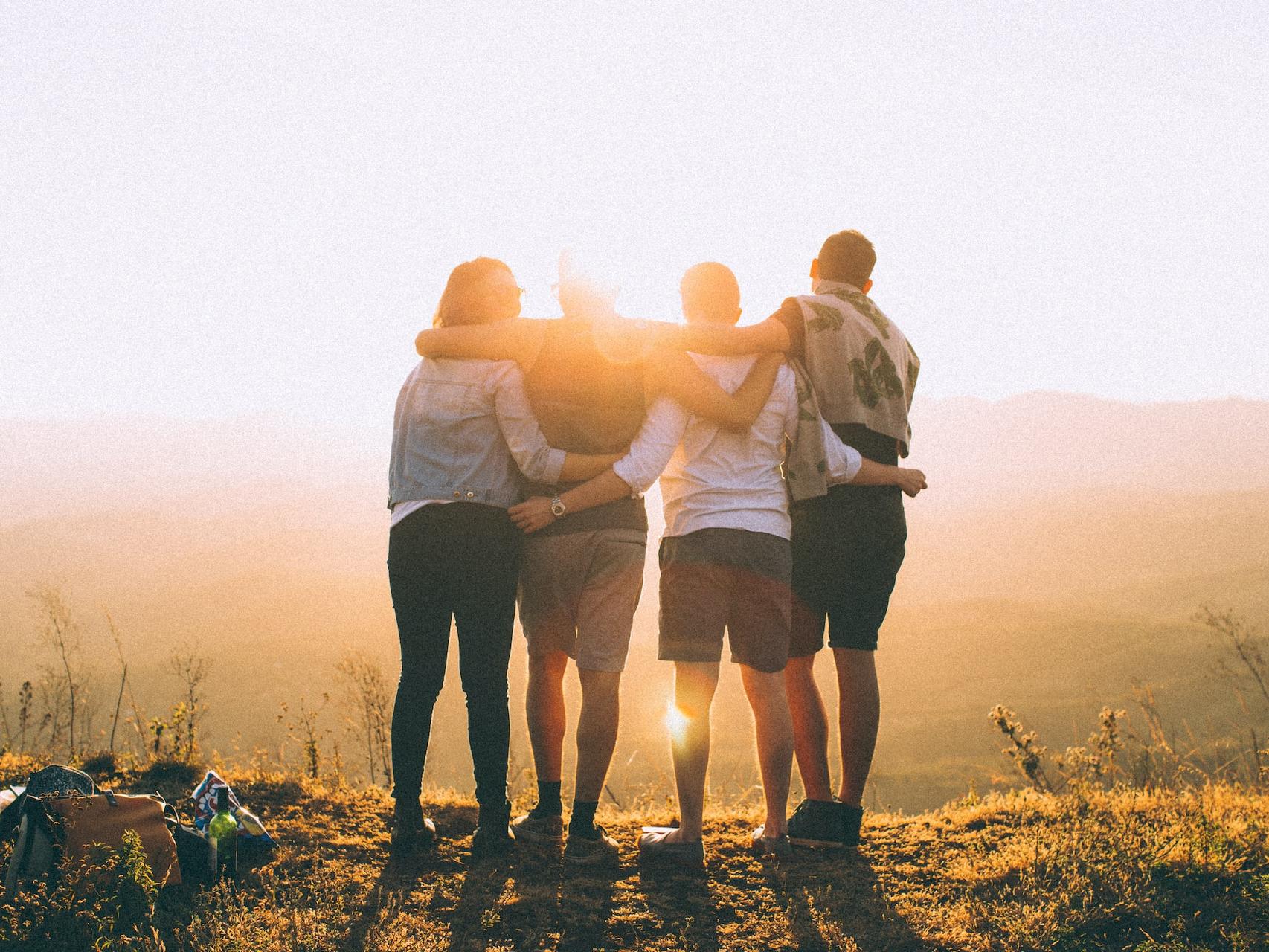 four person hands wrap around shoulders while looking at sunset